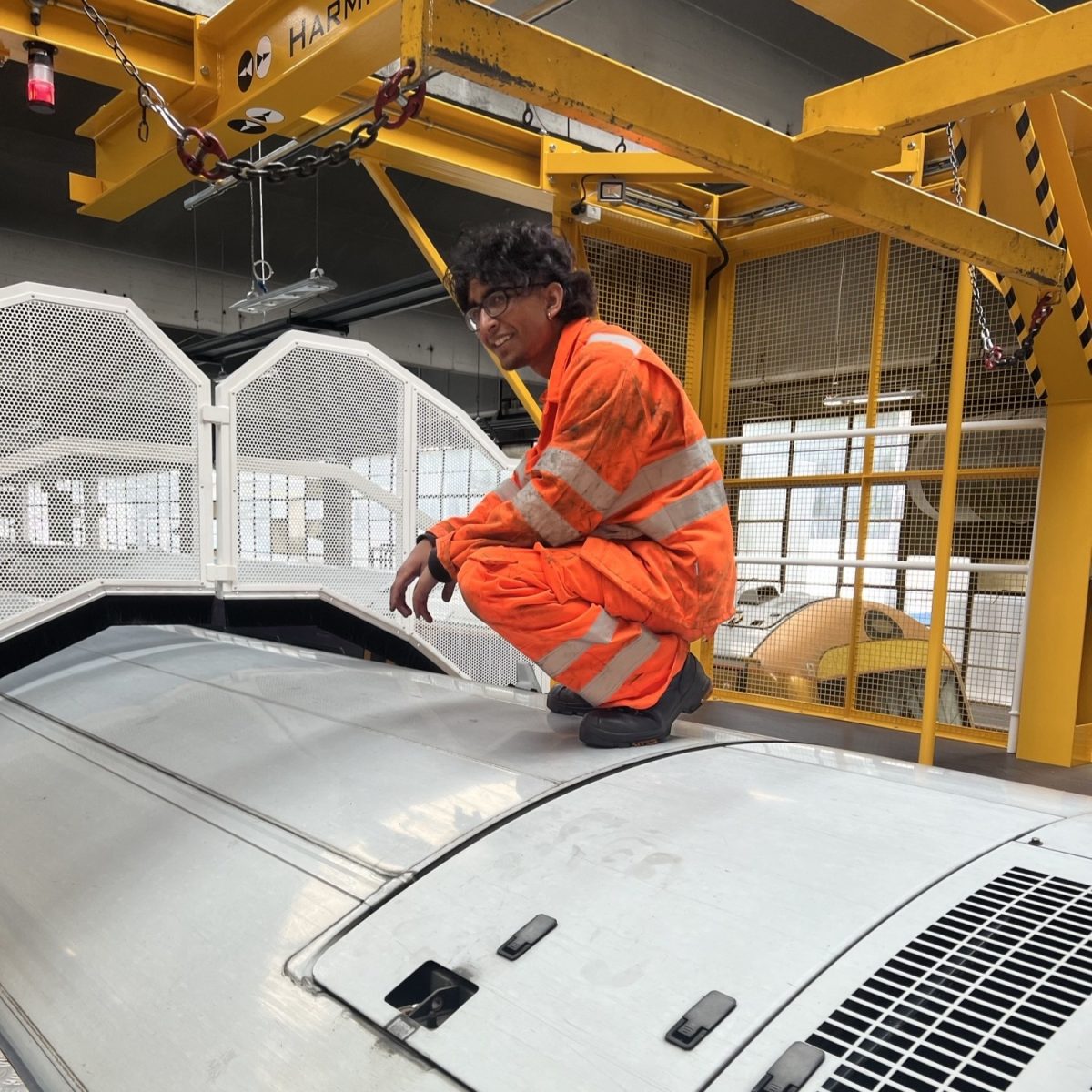 Railway Engineering Technician Dev, wearing orange hi-vis overalls and work boots, crouching on top of a train, in a rail depot workshop.