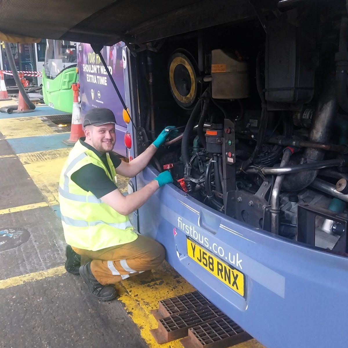 Bus Engineering Technician Kian, wearing a hi-vis vest, gloves, and a cap, working on the engine compartment of a bus, in a depot.