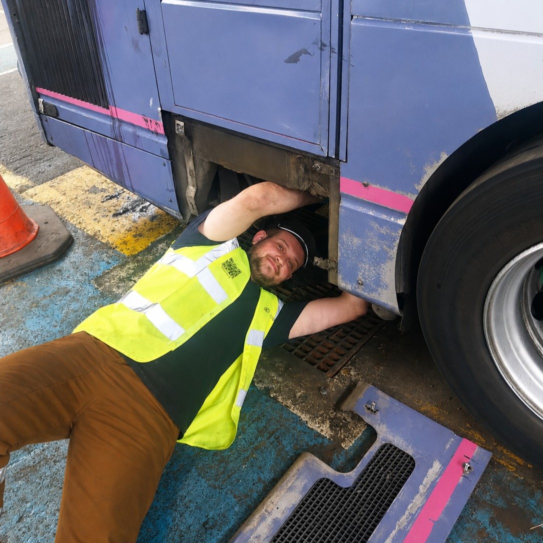 Bus Engineering Technician Kian, wearing a hi-vis vest and cap, lying underneath a bus while reaching into an access panel, in a bus depot.