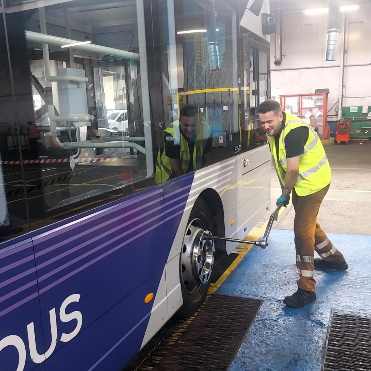 Bus Engineering Technician Kian, wearing a hi-vis vest and gloves, using a long-handled tool on a bus wheel, in a bus depot workshop.