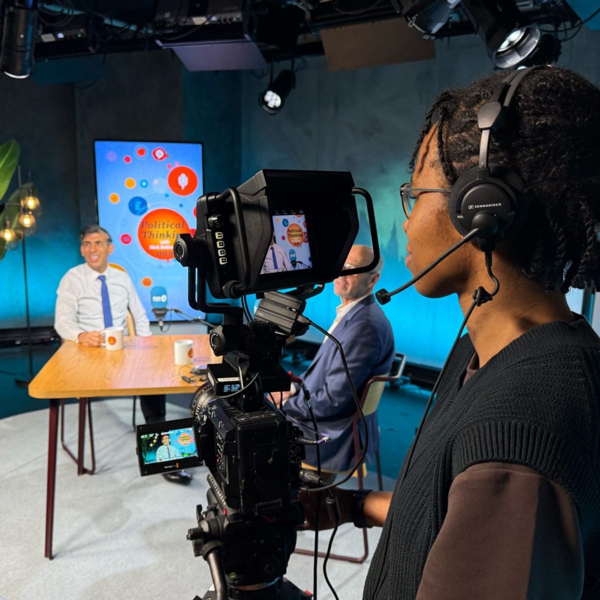 Broadcast technician Maci-Skye, wearing a headset, operating a camera and filming Rishi Sunak being interviewed in a TV studio.
