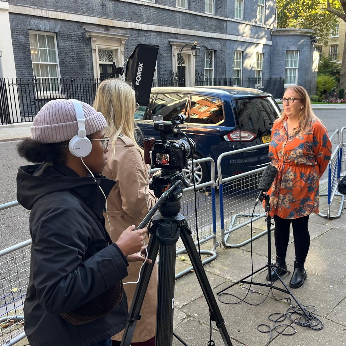 Broadcast technician Maci-Skye, wearing headphones, operating a camera and filming a person being interviewed, outside number 10 Downing Street.