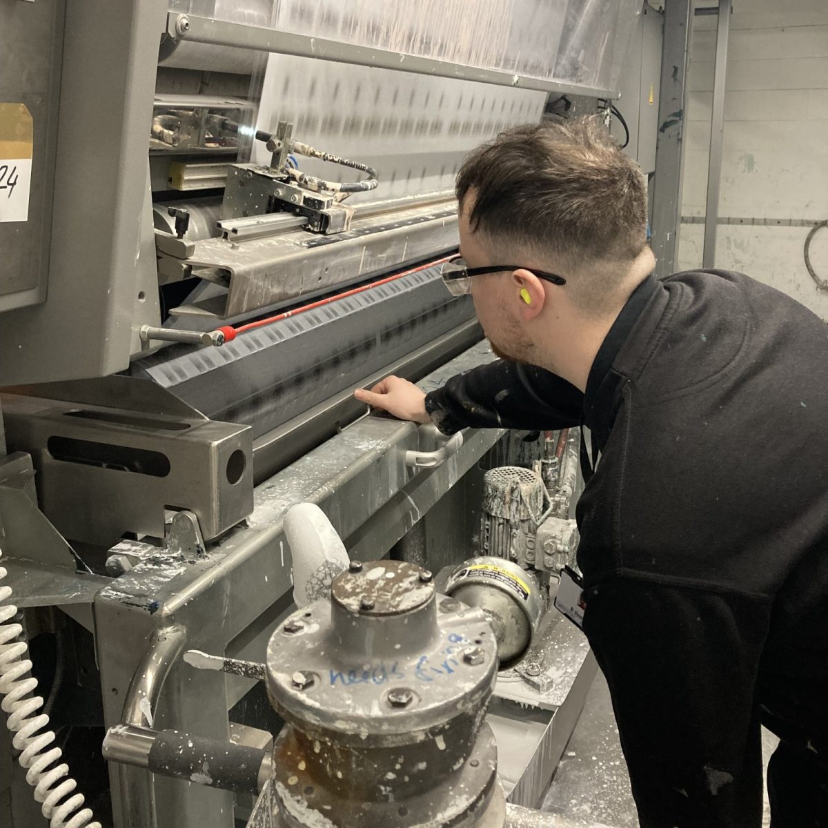 Print Technician Ryan, wearing safety glasses and ear plugs, inspecting the moving parts of an industrial machine, in a manufacturing workshop.