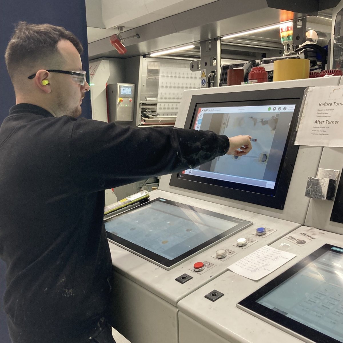 Print Technician Ryan, wearing safety glasses and ear plugs, using a touchscreen control panel to operate an industrial machine, in a manufacturing workshop.