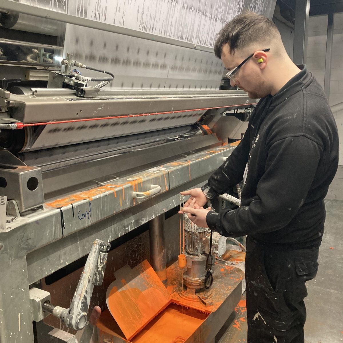 Print Technician Ryan, wearing safety glasses and ear plugs, standing beside a large industrial machine, in a manufacturing workshop, with splashes of orange dye covering the machine and drip tray.