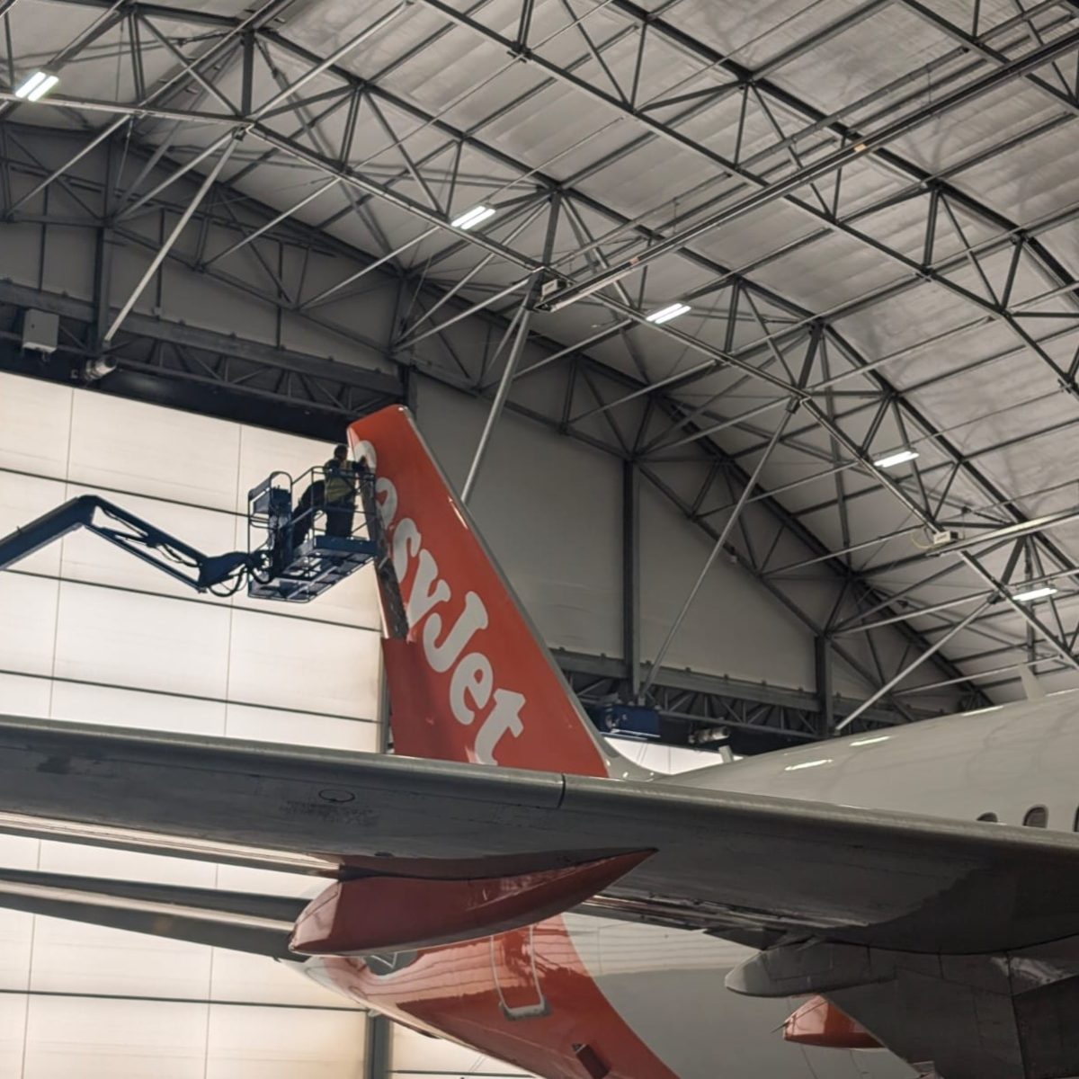Aircraft maintenance technician George, standing on a craned platform, lifted up to reach the tip of an aircraft's tail.