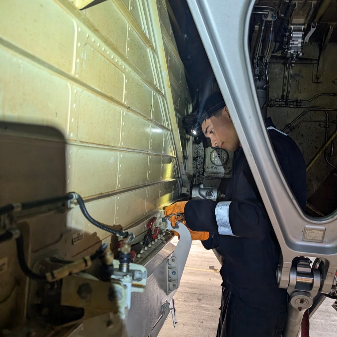 Aircraft maintenance technician George, wearing overalls, gloves, and a headtorch, making adjustments within the landing gear compartment of an aircraft.