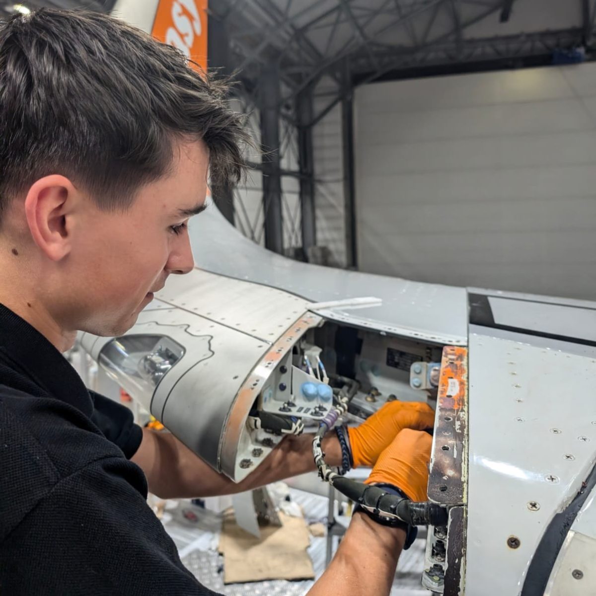 Aircraft maintenance technician George, wearing gloves, working on an open panel of an aircraft's tail, making adjustments to wiring.