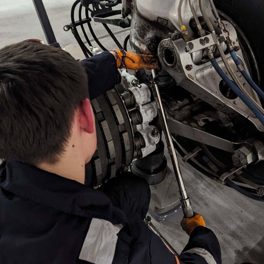 Aircraft maintenance technician George, wearing overalls and gloves, using a long torque wrench to make adjustments to a hydraulic system on an aircraft.