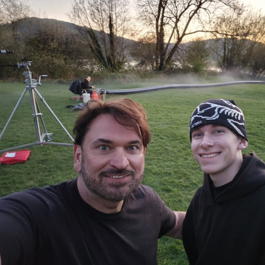 Special Effects Technician Griff and another man during an outside shoot, standing in a field, smiling and posing for the photo, with equipment and a mist machine in the background.