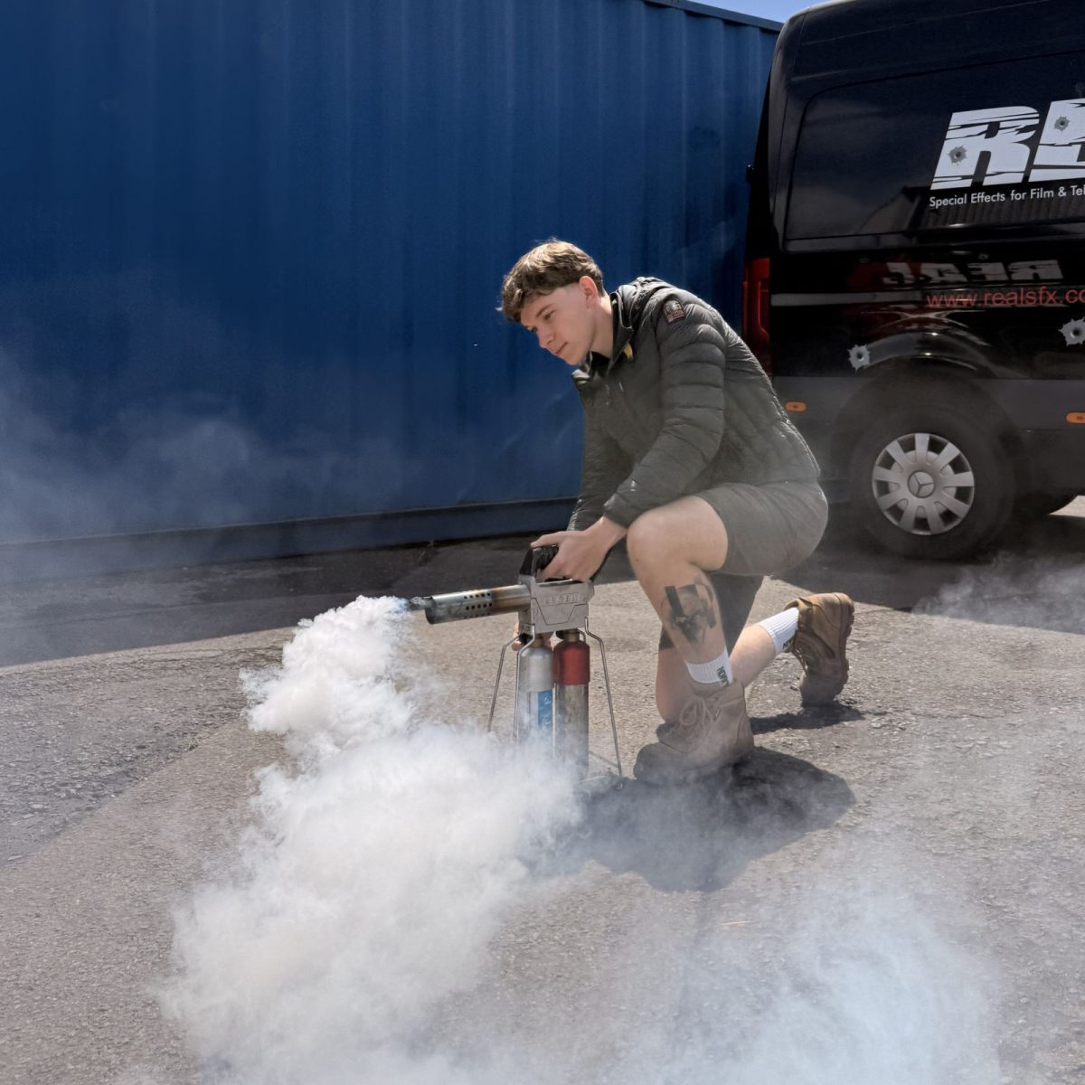 Special Effects Technician Griff, kneeling in a car park, testing a smoke machine, surrounded by smoke.