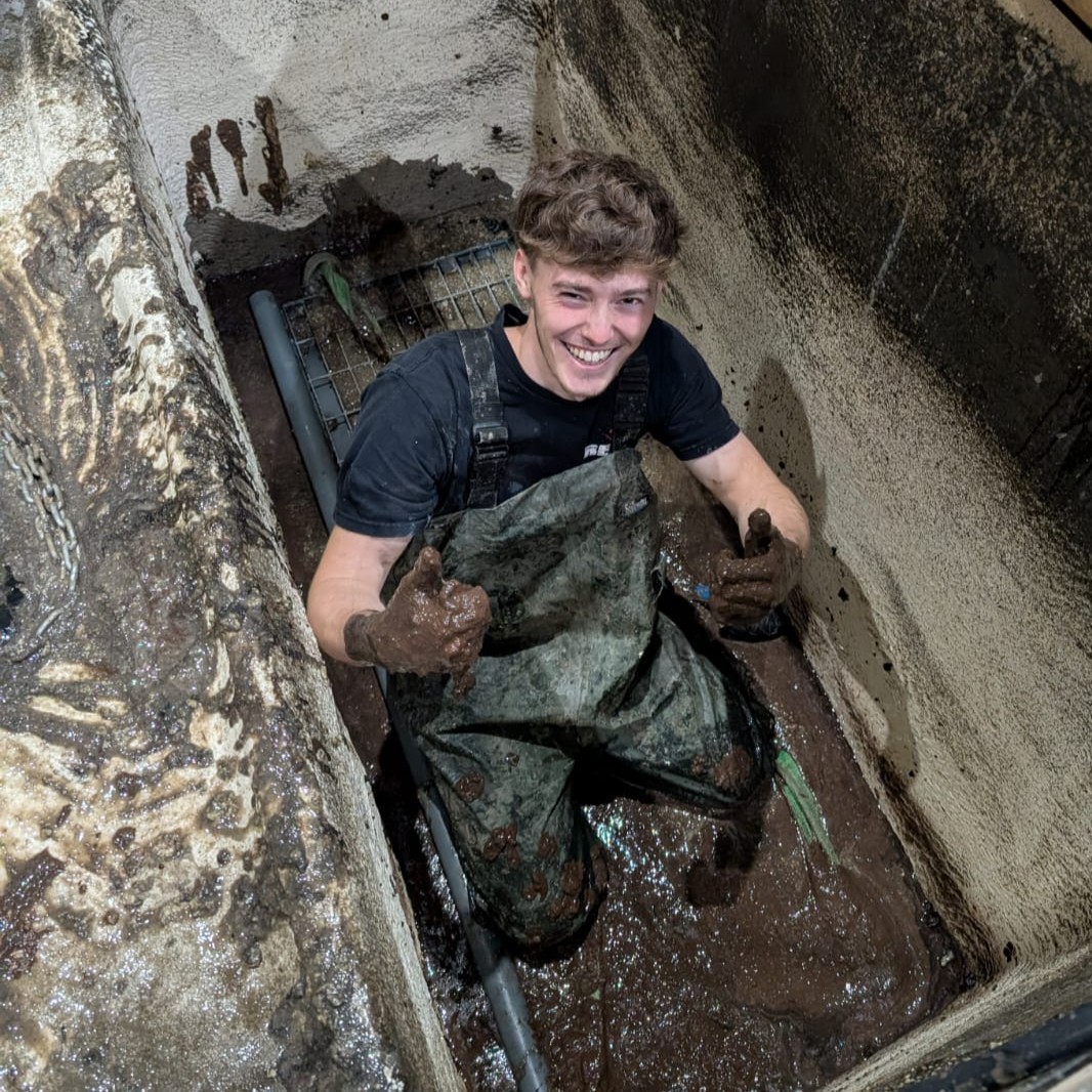 Special Effects Technician Lenny, wearing waders and gloves, in a pit in the floor, covered in mud, smiling and giving a thumbs up.