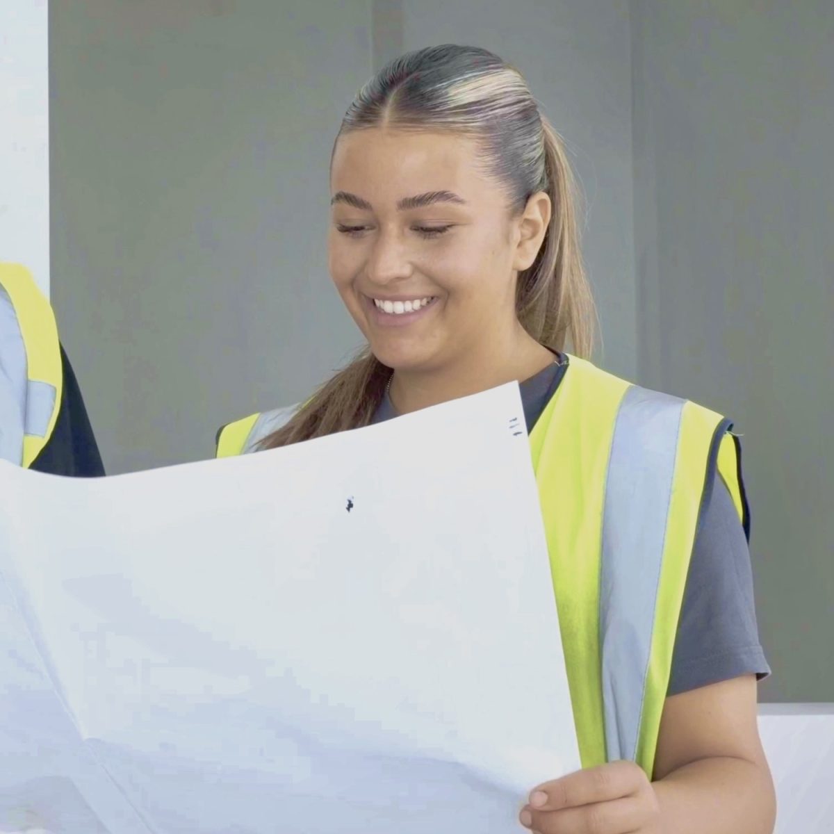Electrician Kelis, wearing a hi-vis vest, smiling, looking at some plans printed on a sheet of paper.