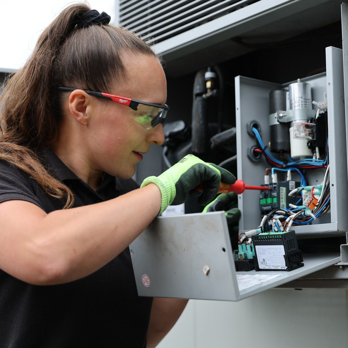 HVAC Technician Chloe, wearing safety glasses and protective gloves, using a screwdriver to work on the electrical components inside an open refrigeration control panel.