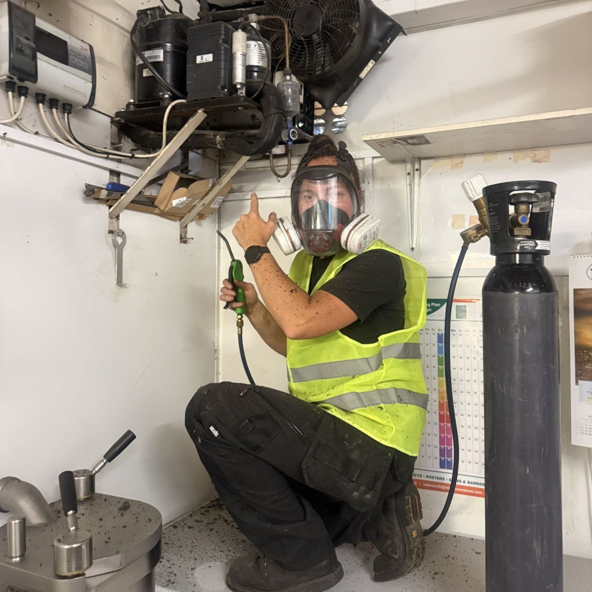 HVAC Technician Chloe wearing a hi-vis vest and respirator mask, holding a hose from a gas canister, kneeling underneath and pointing up to a refrigeration unit mounted near the ceiling.