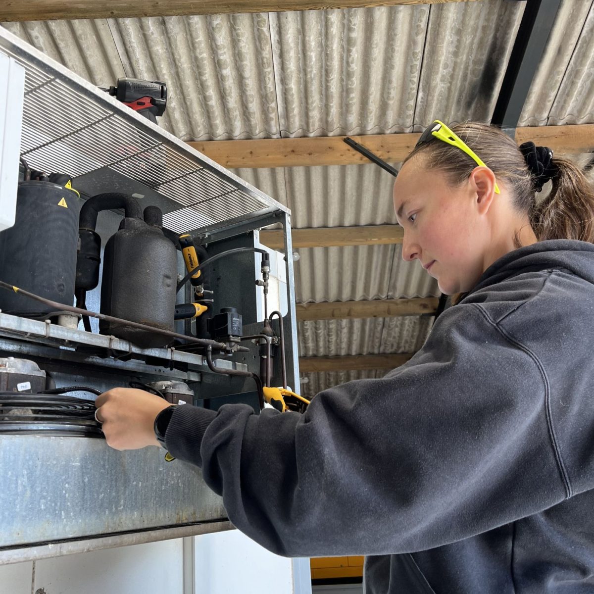 HVAC Technician Chloe, wearing safety glasses on her head, working on the components of a large refrigeration unit, in an industrial workshop.