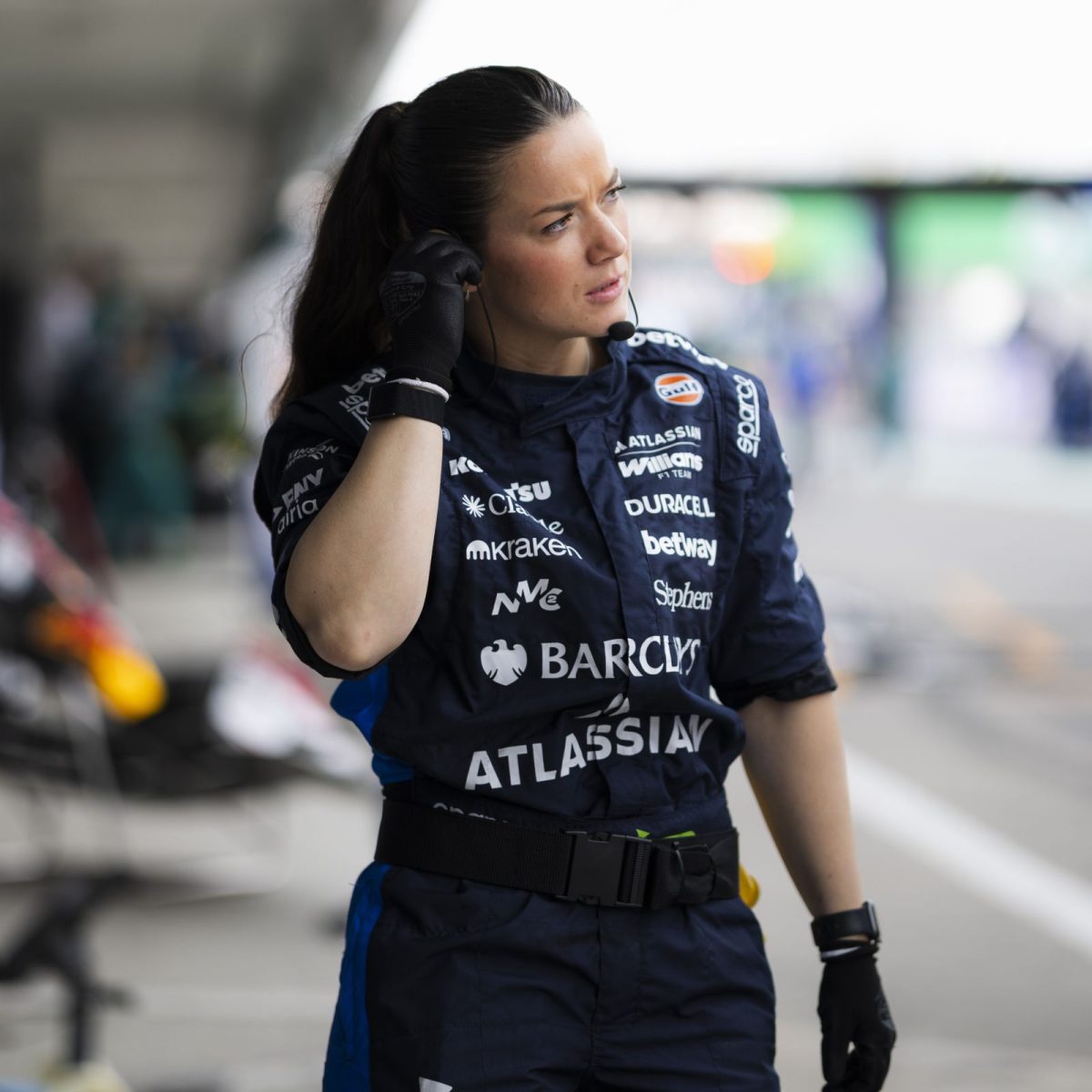 Vehicle Build Technician Cleo, wearing overalls, gloves, and a headset, standing trackside on a race day.