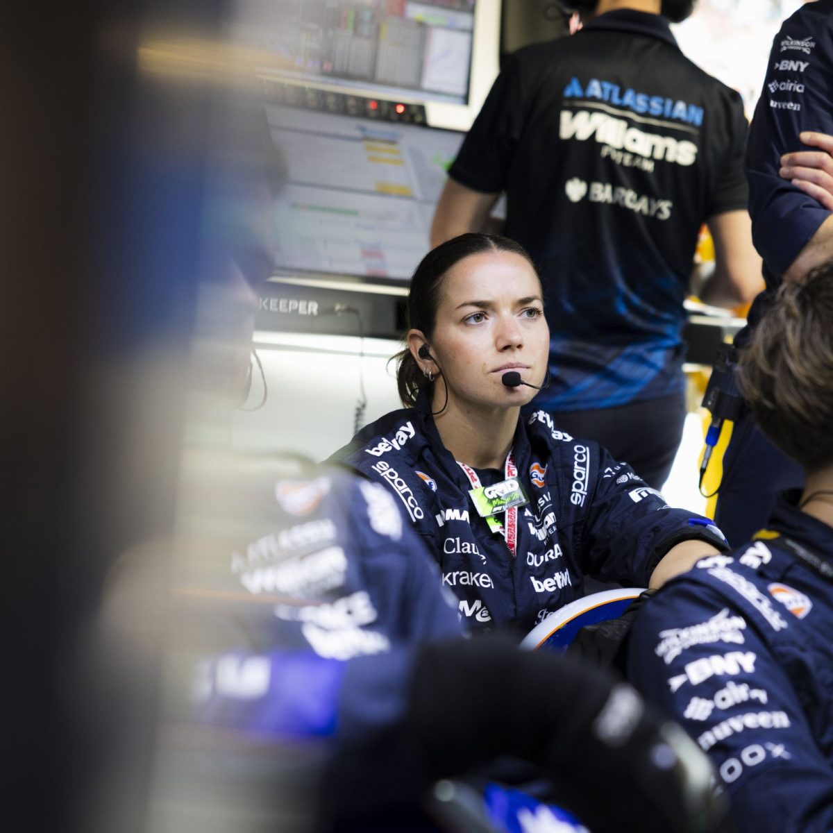 Vehicle Build Technician Cleo, wearing overalls, gloves, and a headset, surrounded by colleagues trackside on a race day, with a focussed look on her face.