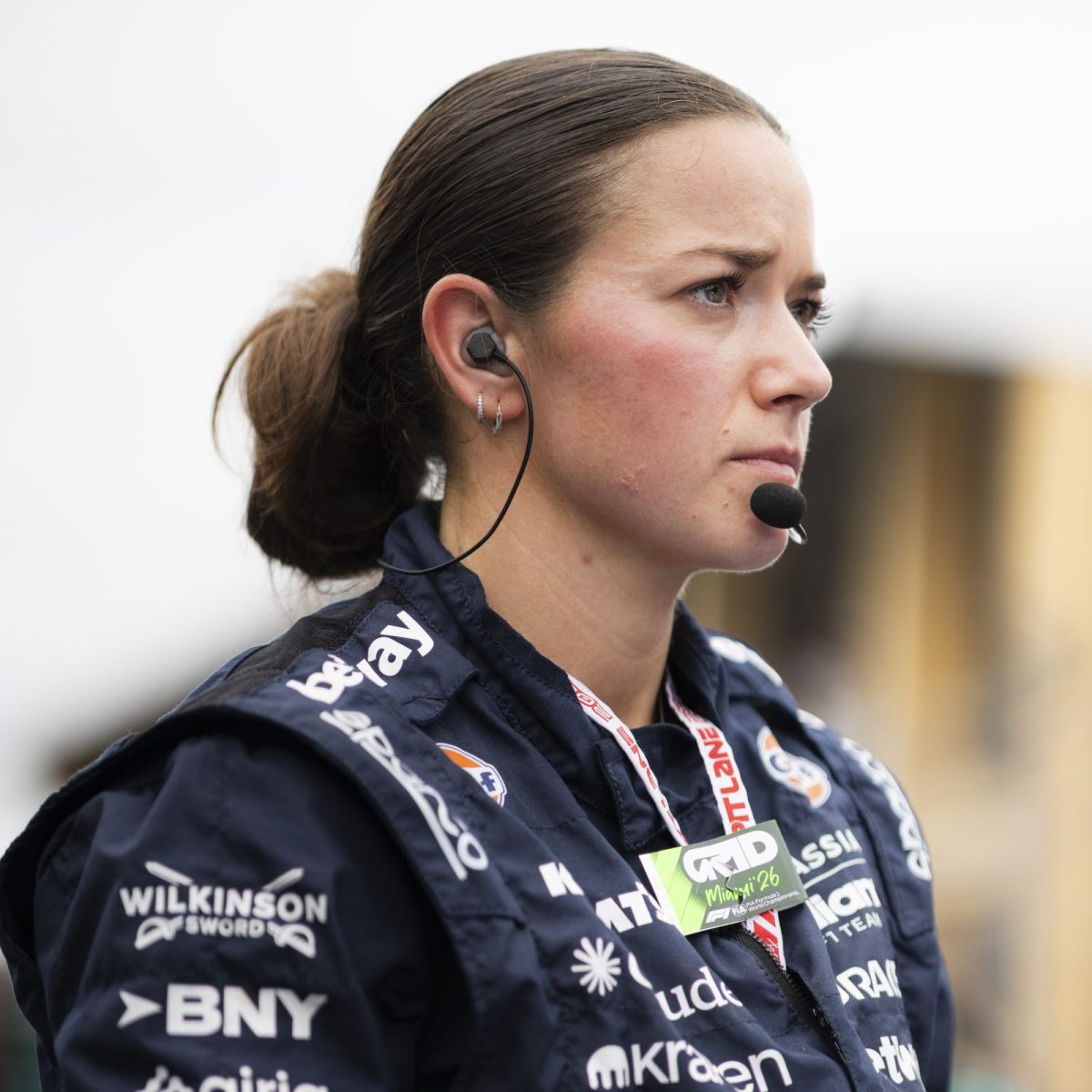 Vehicle Build Technician Cleo, wearing overalls, and a headset, standing trackside on a race day, listening to updates through her earpiece.