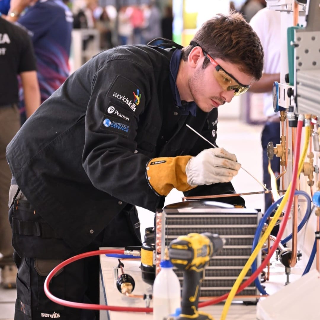 Research and Development Technician Luke, wearing safety glasses and protective gloves, working on a refrigeration unit and copper pipework, in a technical workshop.