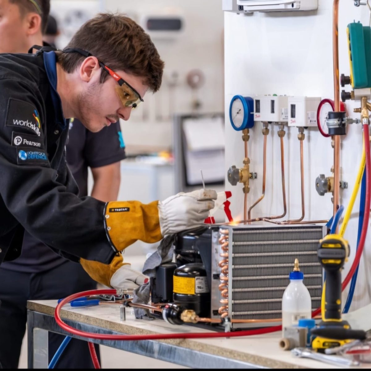 Research and Development Technician Luke, wearing safety glasses and protective gloves, working on a refrigeration unit and copper pipework, in a technical workshop.