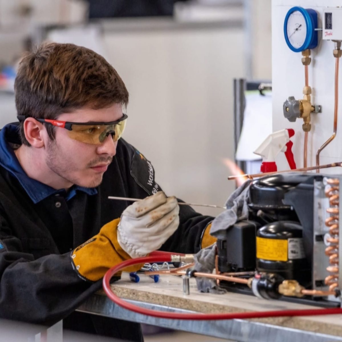 Research and Development Technician Luke, wearing safety glasses and protective gloves, working on a refrigeration unit and copper pipework, in a technical workshop.
